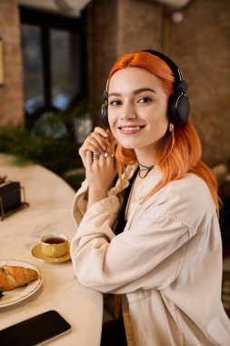 Canlı kızıl saçlı genç bir kadın şık bir kafede kahve içerken müzik dinliyor..