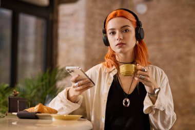 Canlı kızıl saçlı genç bir kadın bir kafede telefonuyla meşgulken kahvesini yudumluyor..