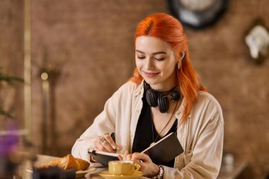 Kızıl saçlı neşeli genç bir kadın bir kafede kahve içerken defterine yazıyor..