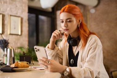 Canlı kızıl saçlı genç bir kadın telefonunu kullanırken şık bir kafede dinleniyor..