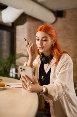 Kızıl saçlı neşeli genç bir kadın bir kafede oturuyor, kahve yudumlarken telefonuyla meşgul..