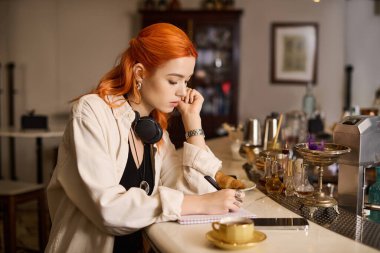 Canlı kızıl saçlı genç bir kadın modern bir kafede notlar alırken kahveyi yudumluyor..