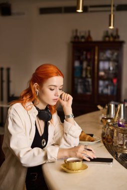 Canlı kızıl saçlı genç bir kadın gündelik bir kafede oturuyor, düşüncelere ve aktivitelere dalmış..