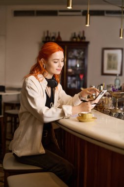 Modern bir kafede canlı kızıl saçlı genç bir kadın rahatlar ve cihazıyla bağlantı kurar..