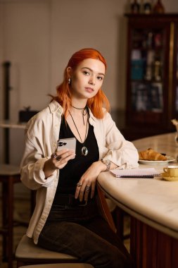 Canlı kızıl saçlı genç bir kadın bir kafede dinleniyor, telefonunu tutarken düşünceli görünüyor..