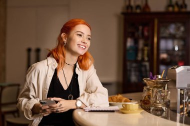 Kızıl saçlı neşeli bir kadın bir kafede oturuyor, gülümsüyor ve kahvesinin ve pastasının tadını çıkarıyor..