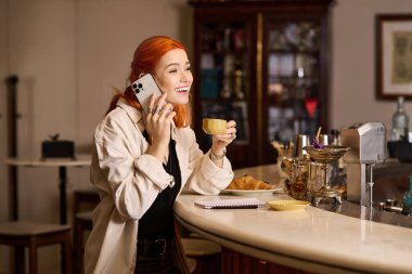 Kızıl saçlı güzel bir kadın gülümsüyor, kahvesini yudumluyor ve telefonda sohbet ediyor..