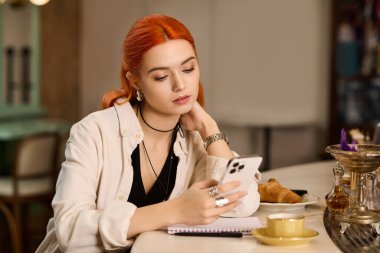Kızıl saçlı genç bir kadın bir kafede dinleniyor, kahvesini yudumluyor ve akıllı telefonunu kullanıyor..