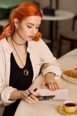 Kızıl saçlı genç bir kadın kahvesini içerken bir kafede notlar yazıyor..