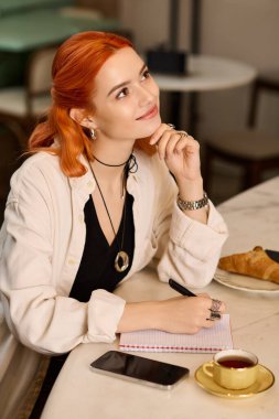 Kızıl saçlı genç bir kadın, hayat dolu bir kafede deftere yazar, kahvesini yudumlar..