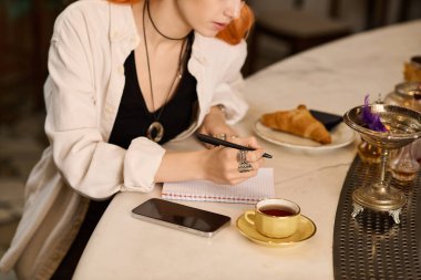 Genç bir kadın defterine bir kafede çay fincanının yanında yazıyor..