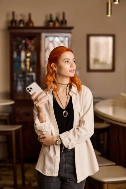 Kızıl saçlı güzel bir kadın canlı bir kafede duruyor, telefonunu tutuyor ve gülümsüyor..