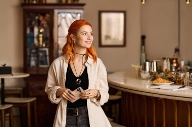 Neşeli genç bir kadın rahat bir kafede duruyor, telefonunu tutuyor ve hafifçe gülümsüyor..