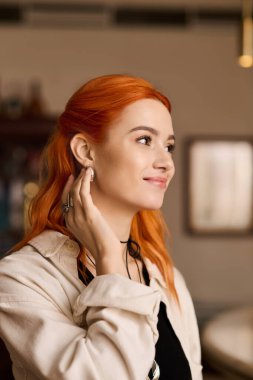 Canlı kızıl saçlı genç bir kadın, rahat bir kafede eğlenirken hafifçe gülümsüyor..