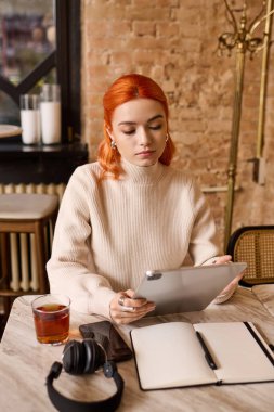 Kızıl saçlı genç bir kadın kafede sıcak bir içecek yudumlarken tabletini kullanıyor..