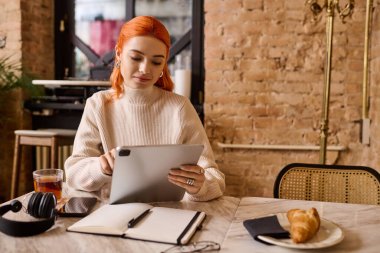 Neşeli kızıl saçlı, mutlu genç bir kadın tabletle çalışıyor, sıcak bir içecek ve kruvasanın tadını çıkarıyor..