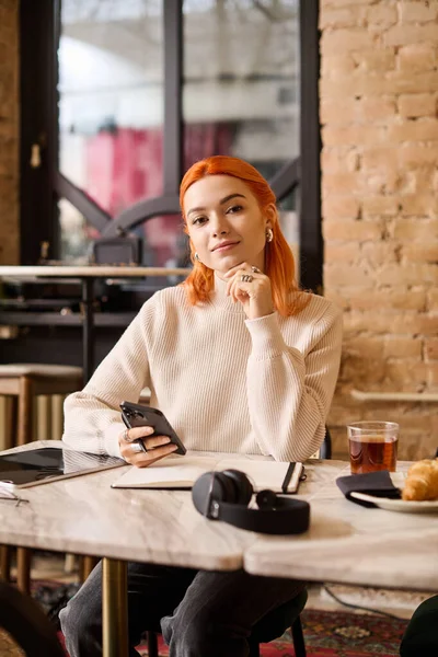 Moda bir kafede, çarpıcı kızıl saçlı genç bir kadın rahat bir şekilde oturur.