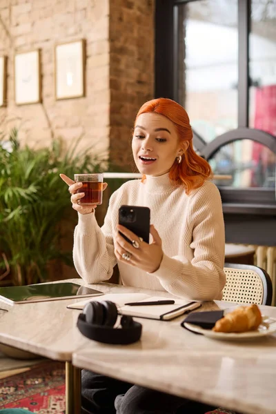Canlı kızıl saçlı genç bir kadın bir kafede akıllı telefonunu kullanırken kahveden zevk alır..