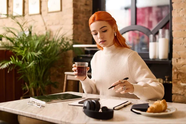 Kızıl saçlı güzel bir kadın, rahat bir kafede yazarken içkisini yudumluyor..