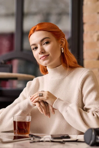 Kızıl saçlı genç bir kadın modern bir kafede rahatlıyor, içkisini yudumluyor ve atmosferin tadını çıkarıyor..