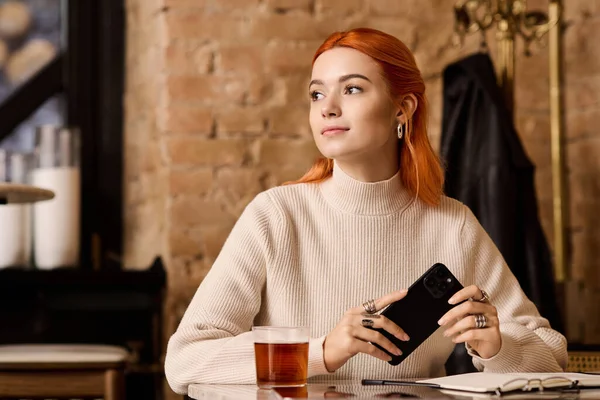 Kızıl saçlı güzel bir kadın modern bir kafede oturuyor, telefonu ve çayıyla rahatlıyor..