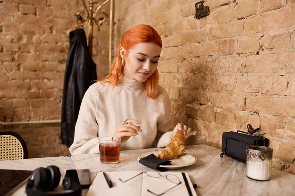 Kızıl saçlı genç bir kadın kafeteryada otururken kruvasan ve çayın tadını çıkarıyor..