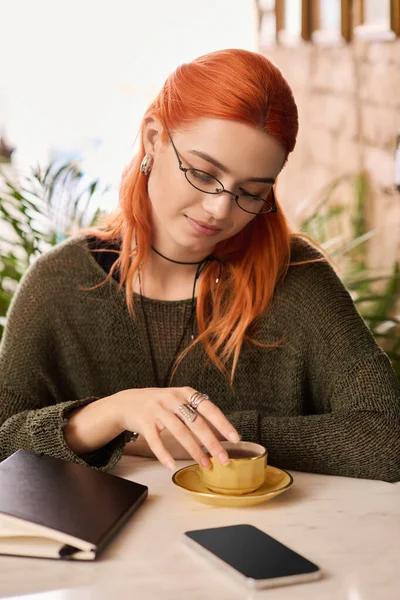 Kızıl saçlı genç bir kadın modern bir kafe ortamında kitap okurken sıcak bir içki içiyor..