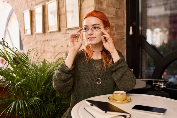Kızıl saçlı genç bir kadın bir kafe masasında oturur, kahvesini yudumlar ve fikir üretir..