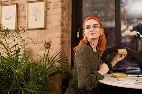 Çarpıcı kızıl saçlı genç bir kadın modern bir kafede rahatlıyor, içkisini yudumluyor ve gülümsüyor..