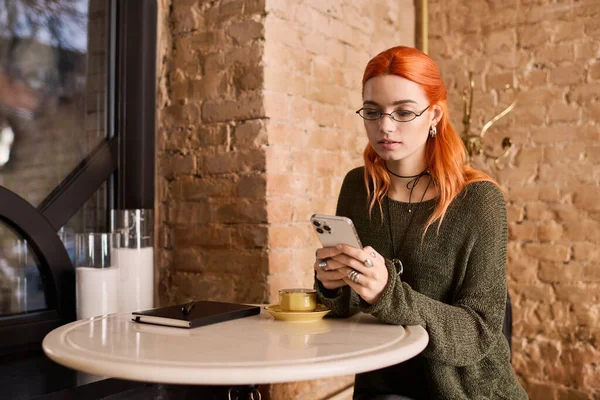 Canlı bir kızıl saçlı şık bir kafede oturur, kahvesini yudumlar ve akıllı telefonuna odaklanır..