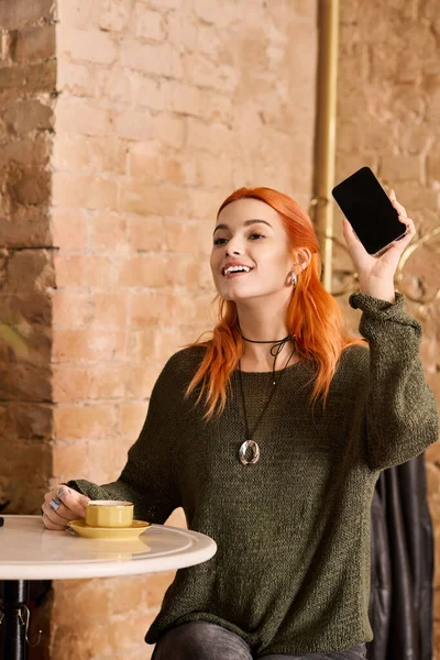Parlak ve mutlu kızıl saçlı kadın, rahat bir kafede kahvesini yudumlarken telefonunu kaldırıyor..