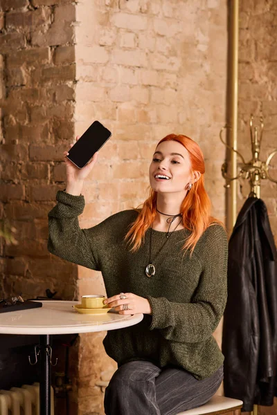 Şık bir kafede kahvesini yudumlarken, kızıl saçlı neşeli bir kadın telefonunu tutuyor..