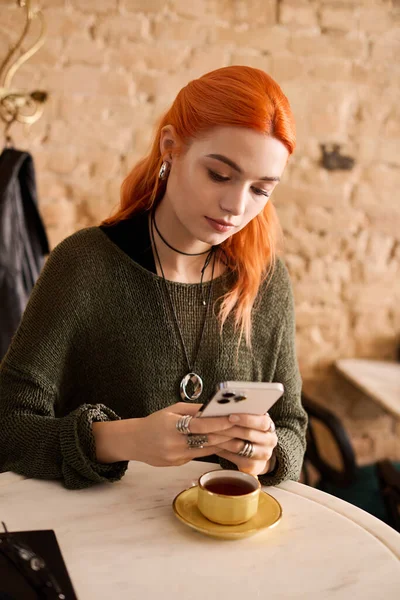 Parlak kızıl saçlı genç bir kadın şık bir kafede dinleniyor, kahvesini yudumluyor ve telefonunu kullanıyor..