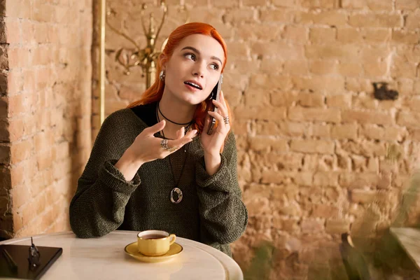 Canlı kızıl saçlı genç bir kadın telefonda sohbet ederken bir kafede rahatlatıcı bir anın tadını çıkarıyor..