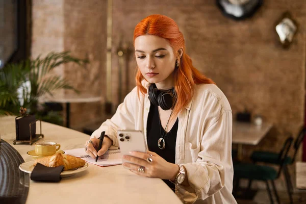 Canlı kızıl saçlı genç bir kadın kafede kahve içerken deftere bir şeyler yazıyor..