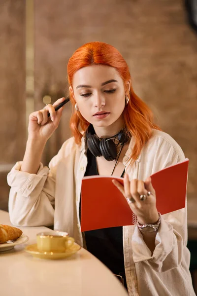 Canlı kızıl saçlı genç bir kadın, modern bir kafede sessiz bir anın tadını çıkarıyor, derinden okumakla meşgul..