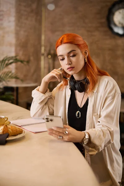 Kızıl saçlı genç bir kadın modern bir kafede oturuyor ve telefonunu incelikle karıştırıyor..