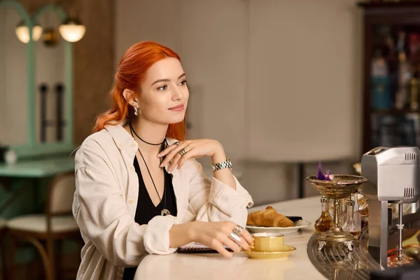 Kızıl saçlı genç bir kadın modern bir kafede dinleniyor, kahvesini yudumluyor ve gülümsüyor..