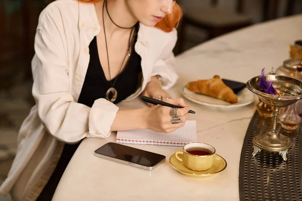Genç bir kadın defterine bir kafede çay fincanının yanında yazıyor..
