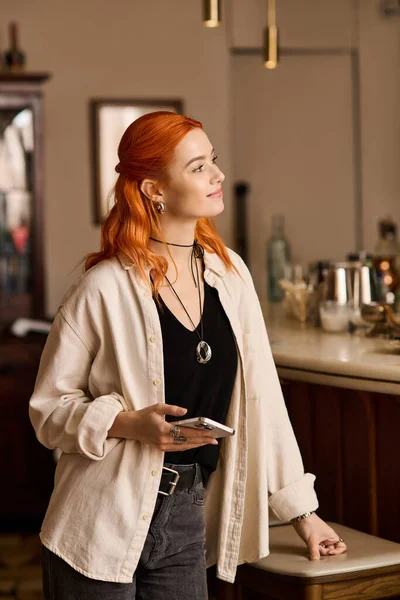 Canlı kızıl saçlı genç bir kadın modern bir kafede dururken hafifçe gülümsüyor..