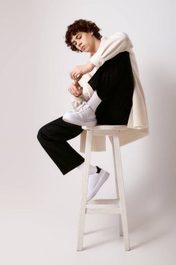 Genç, modaya uygun bir adam bir taburede oturur, rahat ve şık bir görünümü basit bir arka planda sergiler..