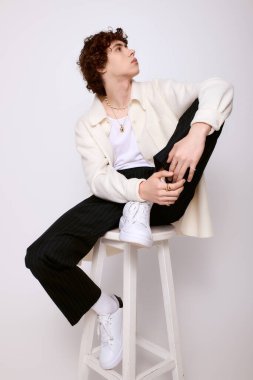 Kıvırcık saçlı genç bir adam bir taburede oturur, şık bir kıyafete güvenir ve stil yayar..