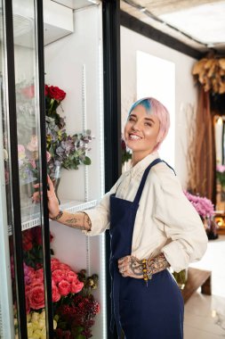 Kısa boyalı saçlı neşeli bir çiçekçi, renkli çiçeklerle çevrili rahat çiçekçisinde çalışıyor..
