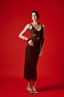 Parlak kahverengi elbiseli kendine güvenen bir kadın kırmızı bir arkaplan karşısında zarafet saçar..