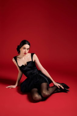 A confident woman in a chic black dress lounges on the floor against a bold red backdrop.