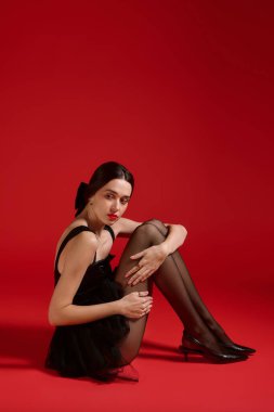 Siyah elbiseli ve kırmızı dudaklı şık bir kadın zarif bir şekilde yerde oturuyor ve bir açıklama yapıyor..