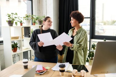 Genç ve cüsseli kadın ve Afrikalı Amerikalı meslektaşı modern bir ofiste belgeleri tartışıyorlar..