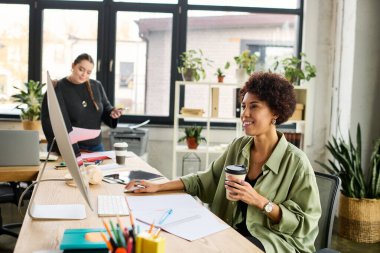 Parlak bir ofiste, iri yarı bir kadın iş arkadaşıyla bir proje üzerinde çalışırken kahve içmekten zevk alır..