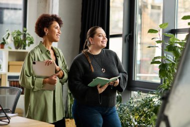 İki kadın yönetici, canlı bir ofis ortamında çalışırken aynı fikirleri paylaşıyor ve gülümsüyor..