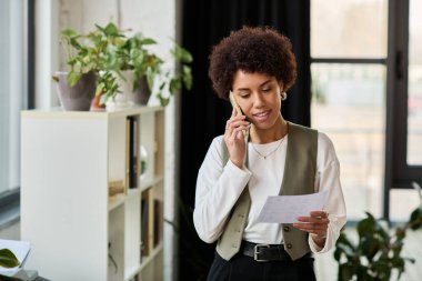 Genç bir Afrikalı Amerikalı kadın, iş yerinde telefonla konuşurken elinde bir belge tutuyor..